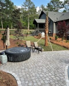 A residential backyard with a paved patio area, outdoor furniture, and landscaped garden leading to a house with a raised deck.