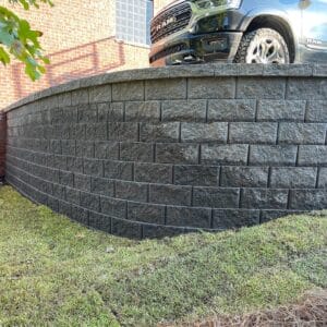 A black retaining wall curves in front of a parked ram truck beside a grassy area and a building with a brick wall.