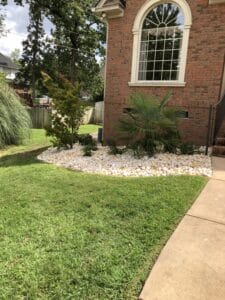 A residential garden bed with white rocks surrounded by green lawn in front of a brick house with a tall arched window.