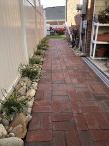 A narrow brick pathway lined with stones and plants leading to a residential courtyard.