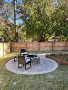 A cozy backyard patio with seating and a fire pit surrounded by a wooden fence and autumn trees.