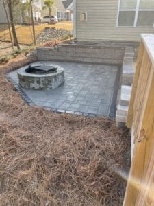 A newly constructed outdoor fire pit area with paving stones and a retaining wall, adjacent to a wooden fence, with construction debris in the surrounding area.