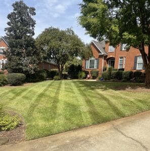 A well-manicured front lawn of a suburban brick house with trees and shrubs on a sunny day.