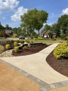 A landscaped suburban area with a winding pathway, manicured shrubs, and trees under a clear sky.