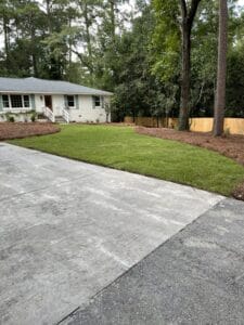 A suburban house with a white exterior, neatly trimmed lawn, and a concrete driveway, surrounded by trees and a wooden fence.