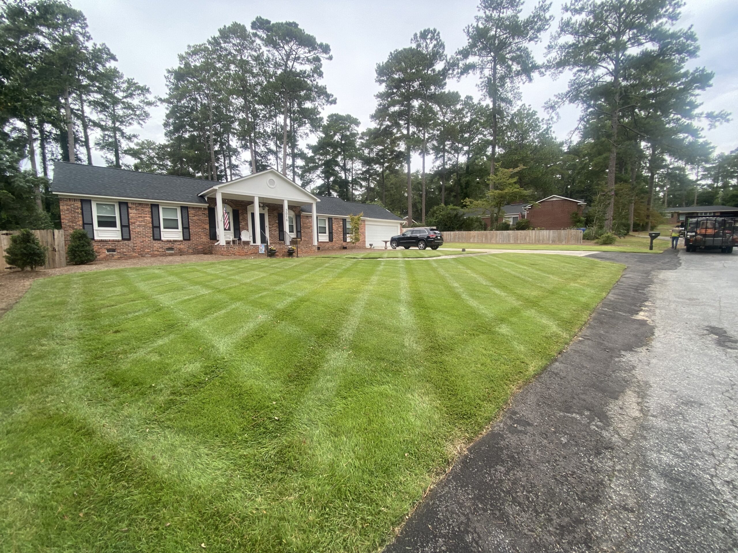 Single-story brick house with white trim, a manicured lawn showing diagonal mowing lines, a driveway, and several parked vehicles; trees line the background.